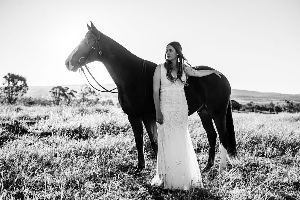 Horse and person in white dress standing together in a sunlit field against rolling hills.