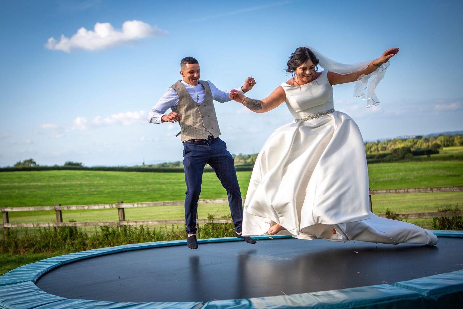 Bride and groom joyfully jumping on a trampoline in wedding attire with green fields in the background.