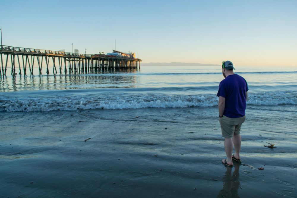Person watching the ocean waves from beach near wooden fishing pier at dawn.