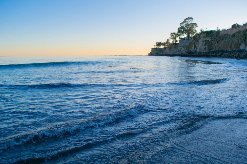Coastal landscape at sunset with blue ocean waves rolling onto shore near rocky cliffs and trees.