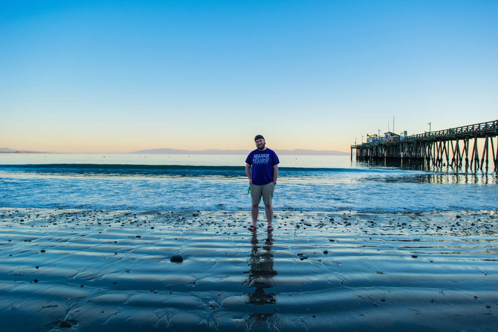 Silhouetted figure stands on wet beach reflecting sunset light near wooden pier.
