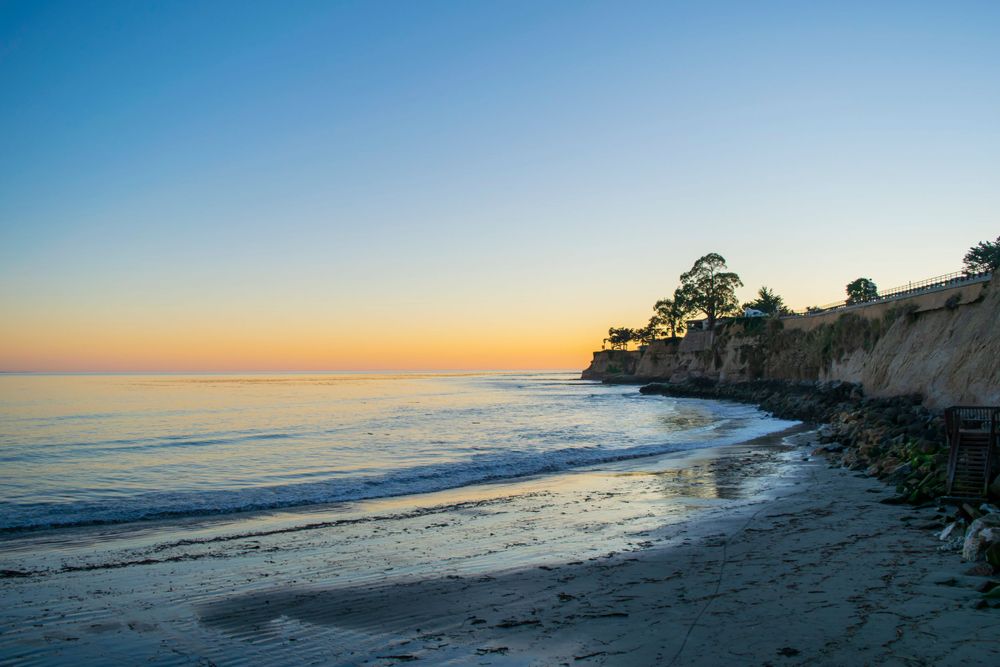 Serene beach landscape at sunset with cliffs and trees overlooking calm ocean waters.