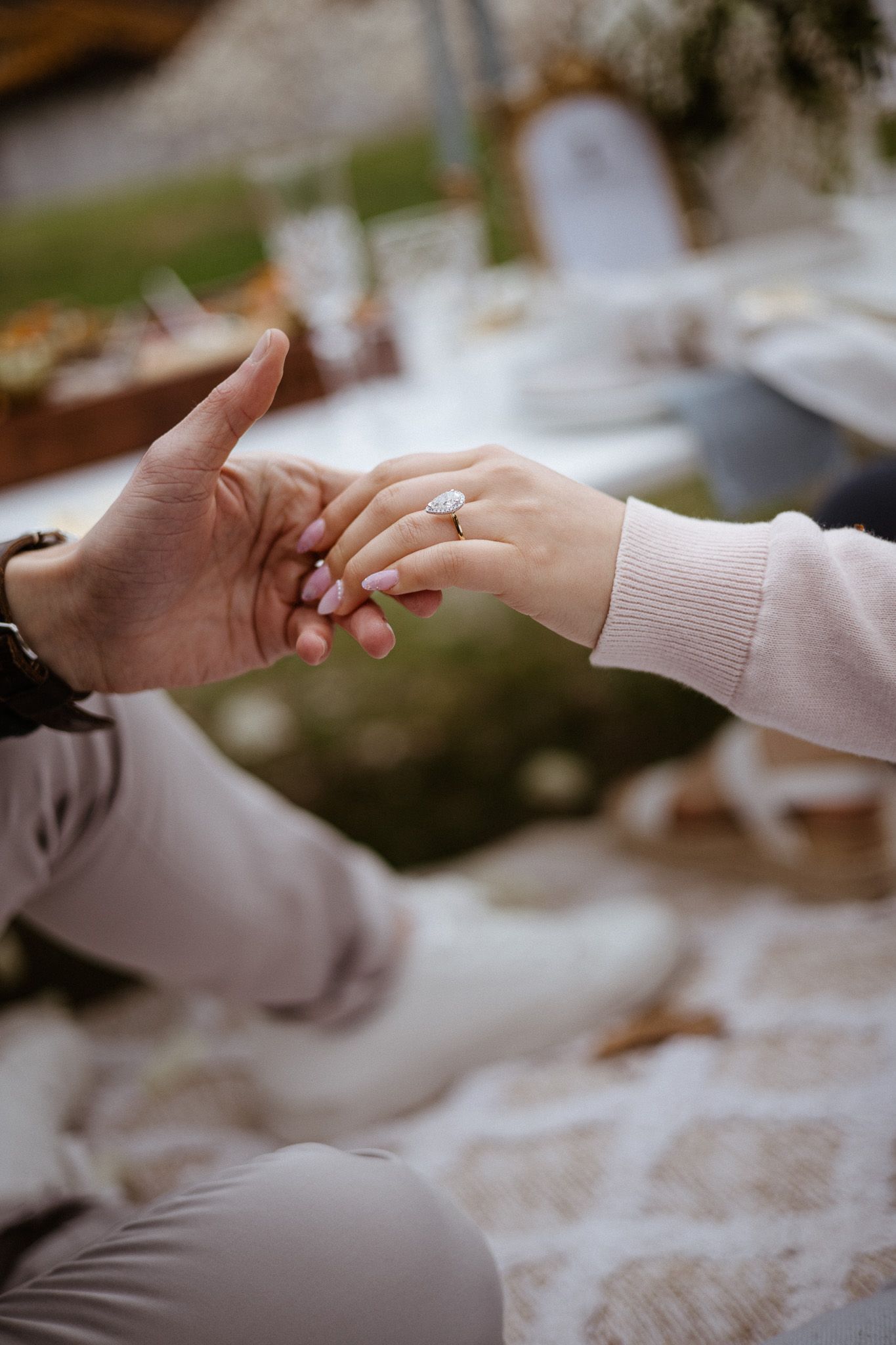 Close-up of hands with engagement ring during romantic picnic moment, one hand wearing light beige sweater.