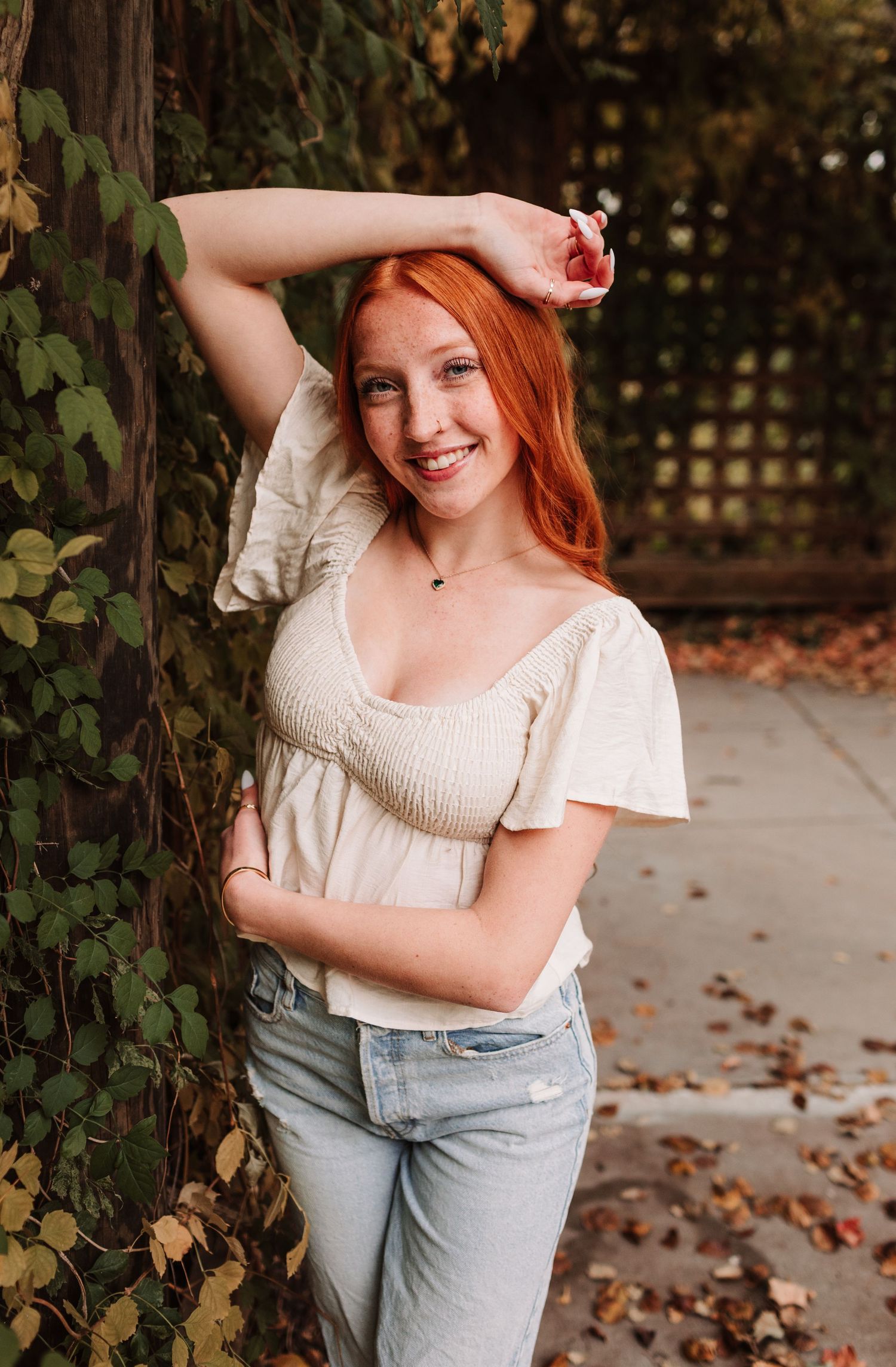 Smiling redhead woman in cream top leaning against ivy-covered wall outdoors.