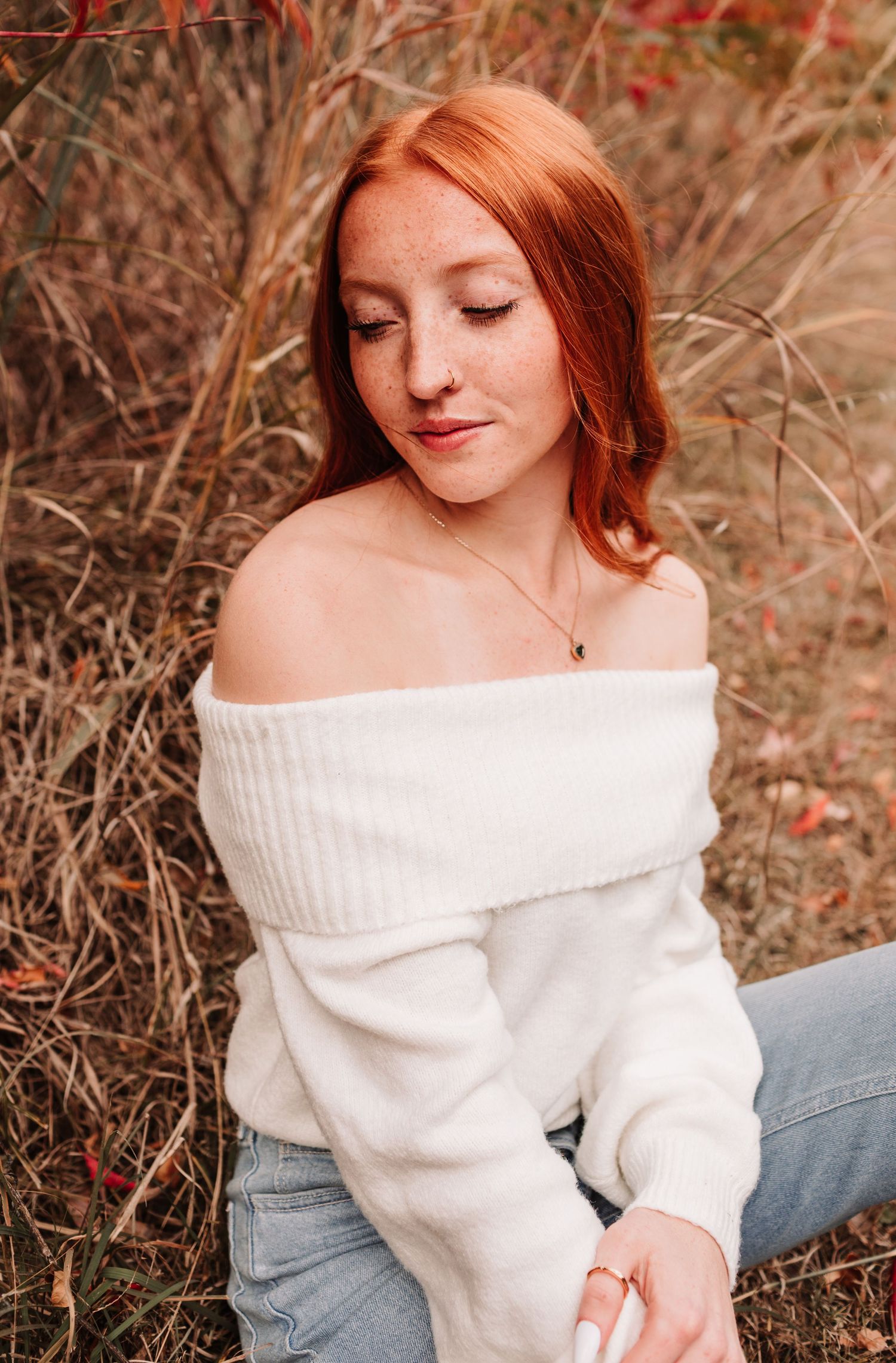 Redhead young woman in off-shoulder white sweater sitting in autumn field.