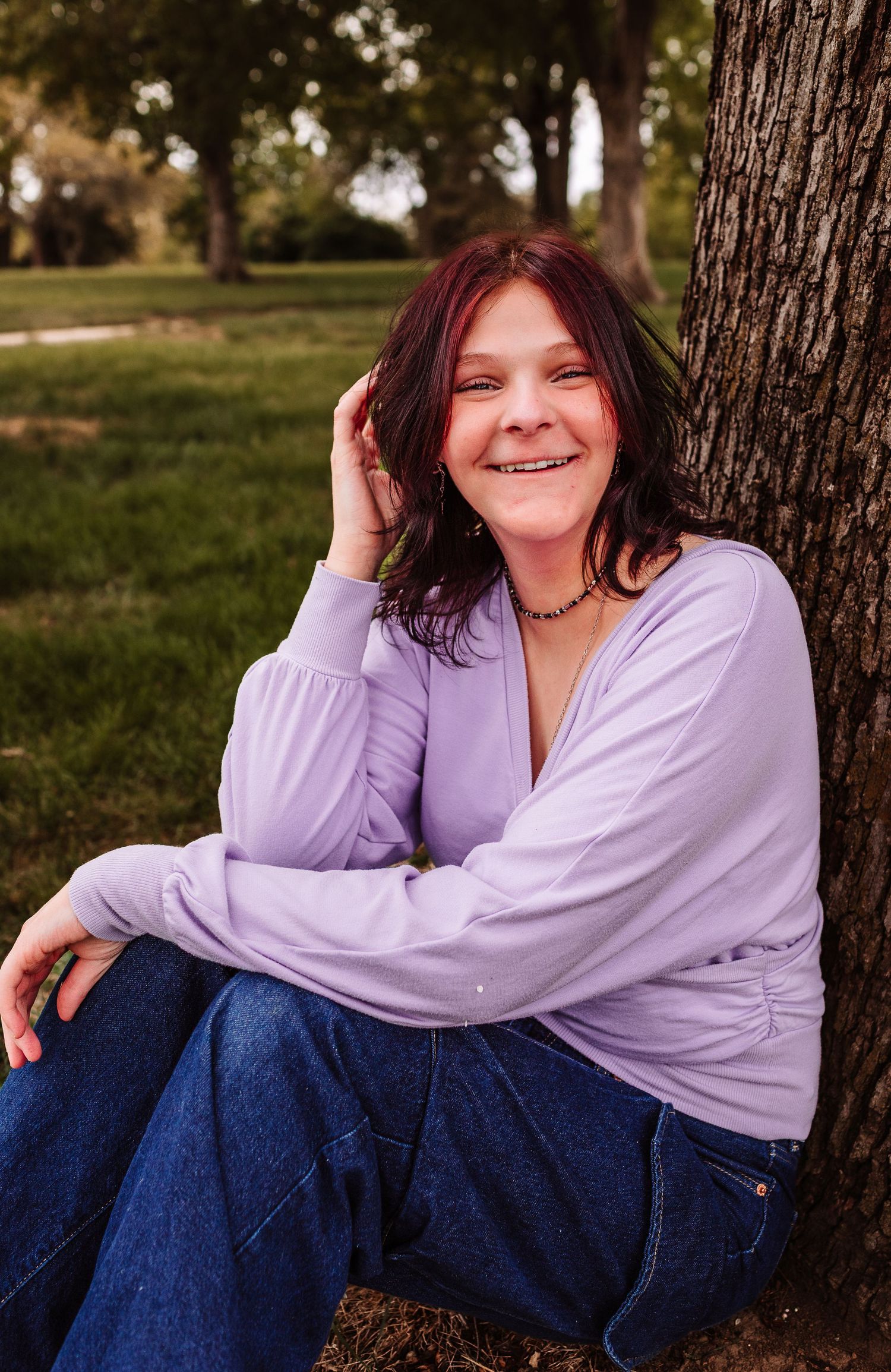 A smiling woman with dark red hair sits against a tree in a park, wearing a lavender top and blue jeans.