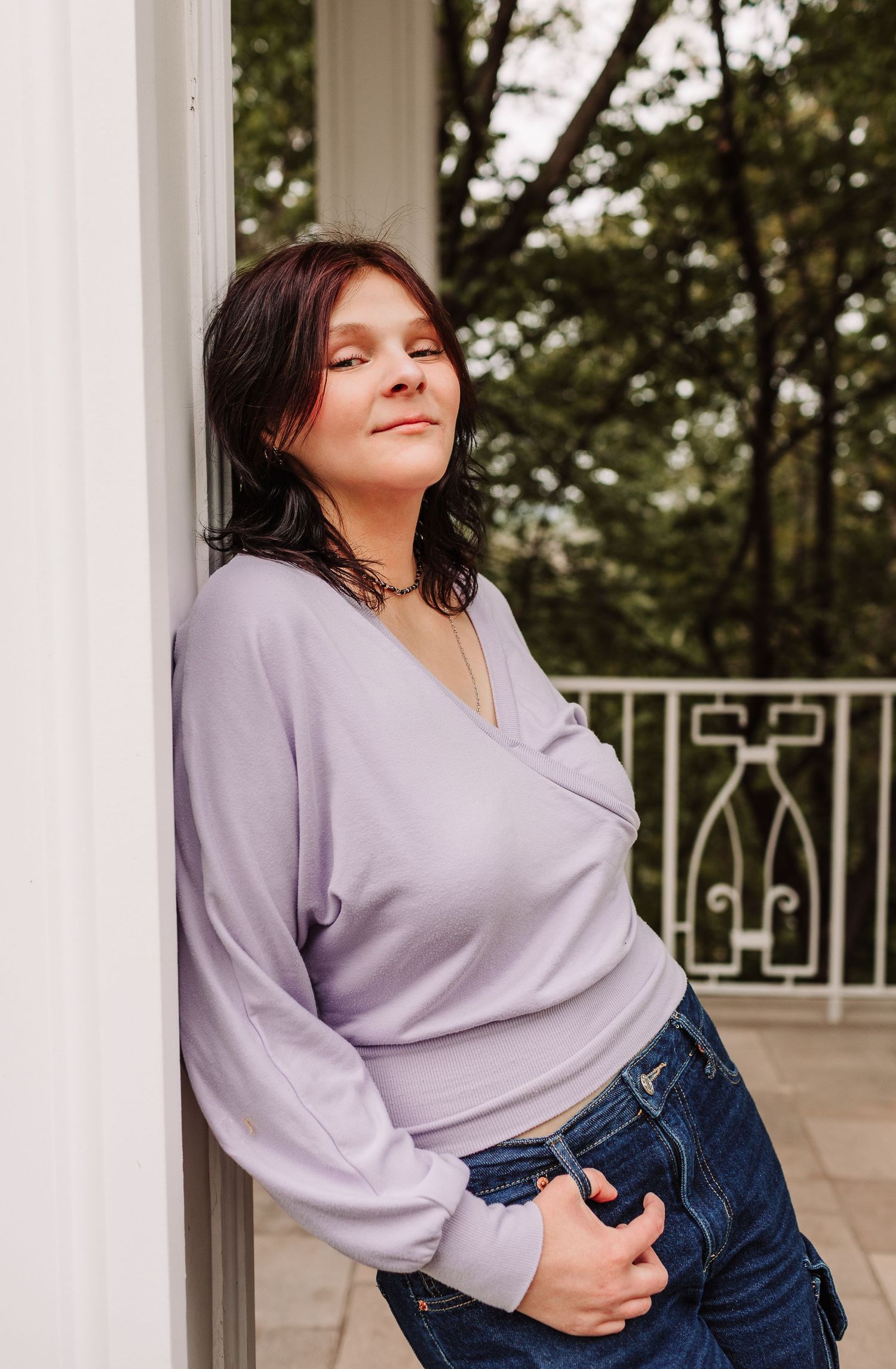 Young woman in lavender wrap top and jeans leaning against white pillar on outdoor balcony with trees in background.