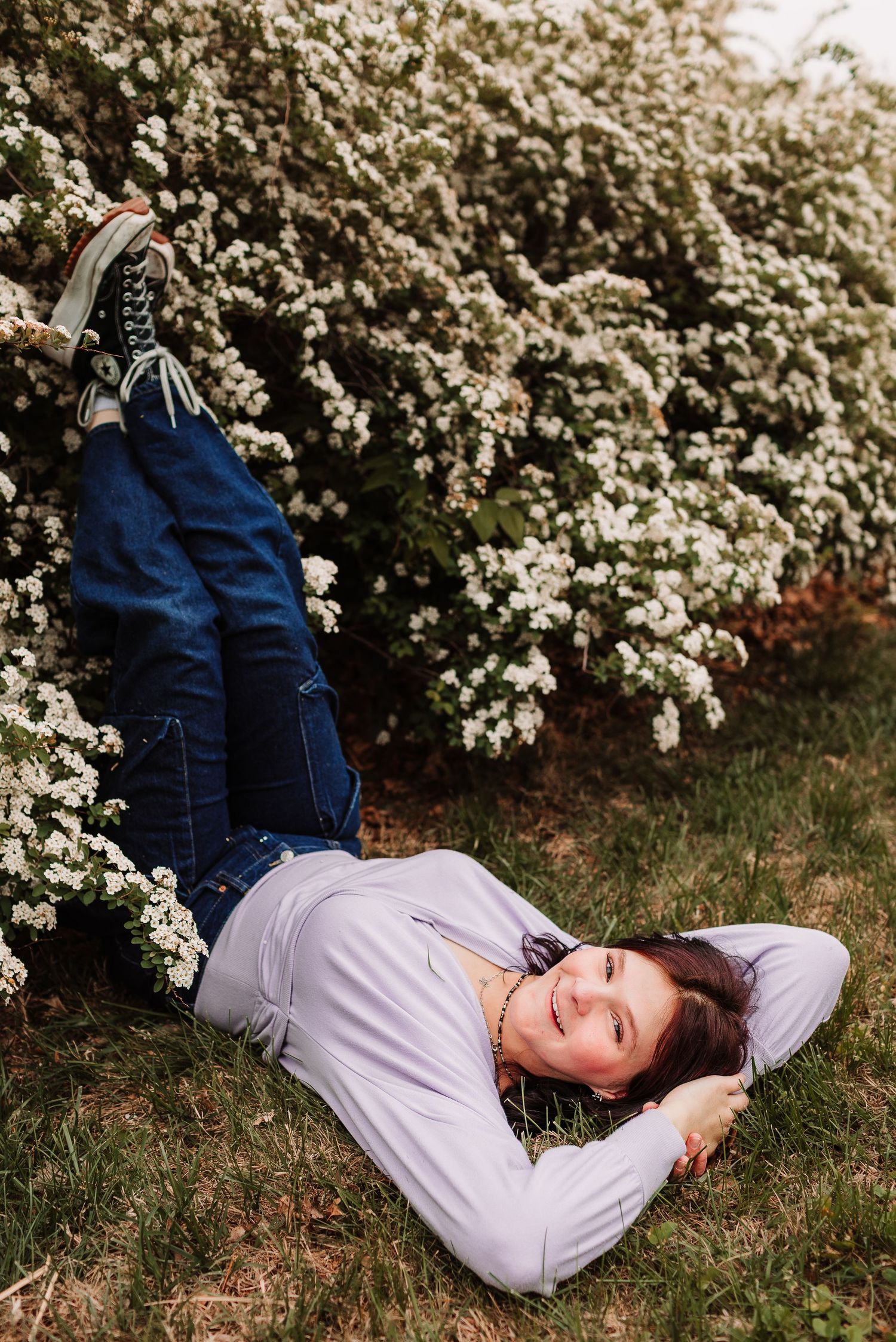Young woman in lavender top and jeans lying on grass smiling beside white flowering bushes outdoors.