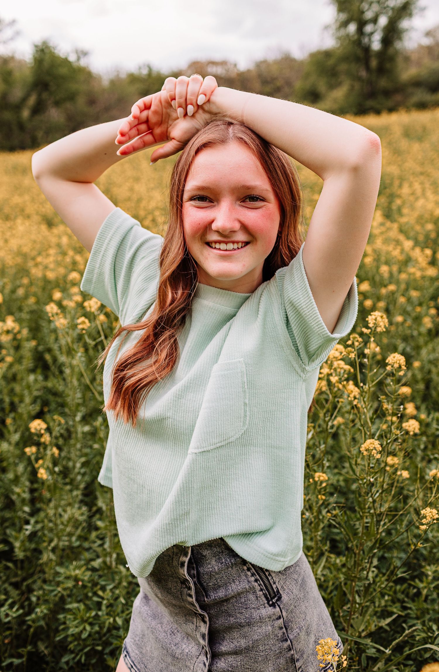 Smiling young woman with red hair in mint green tee poses in vibrant yellow wildflower field outdoors.