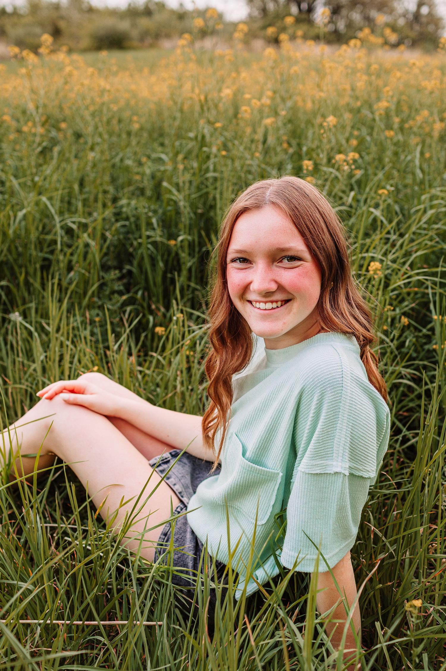 Young woman with red hair smiling in a field of green grass and yellow wildflowers, wearing a mint top and denim shorts.