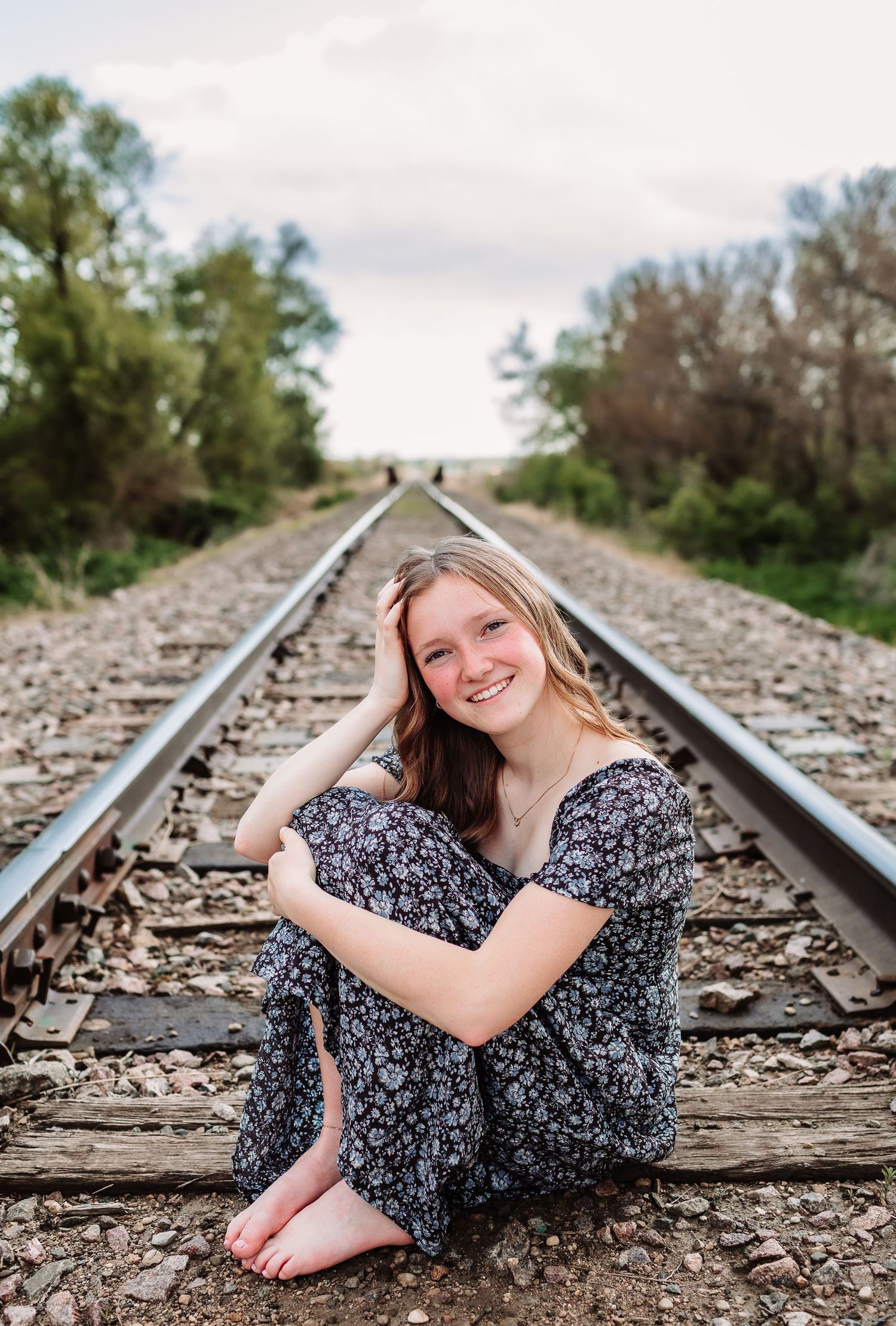 Smiling teenage girl in floral dress sitting barefoot on railroad tracks surrounded by trees.
