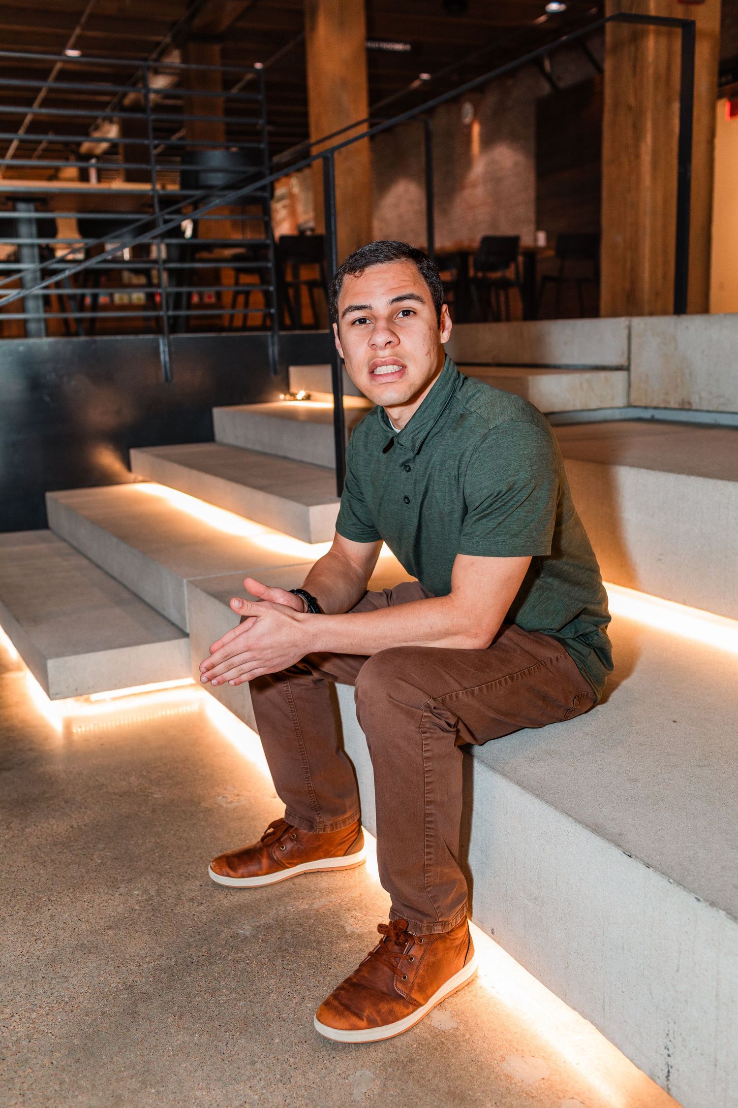 Young man in green polo sitting on illuminated concrete steps in modern venue.