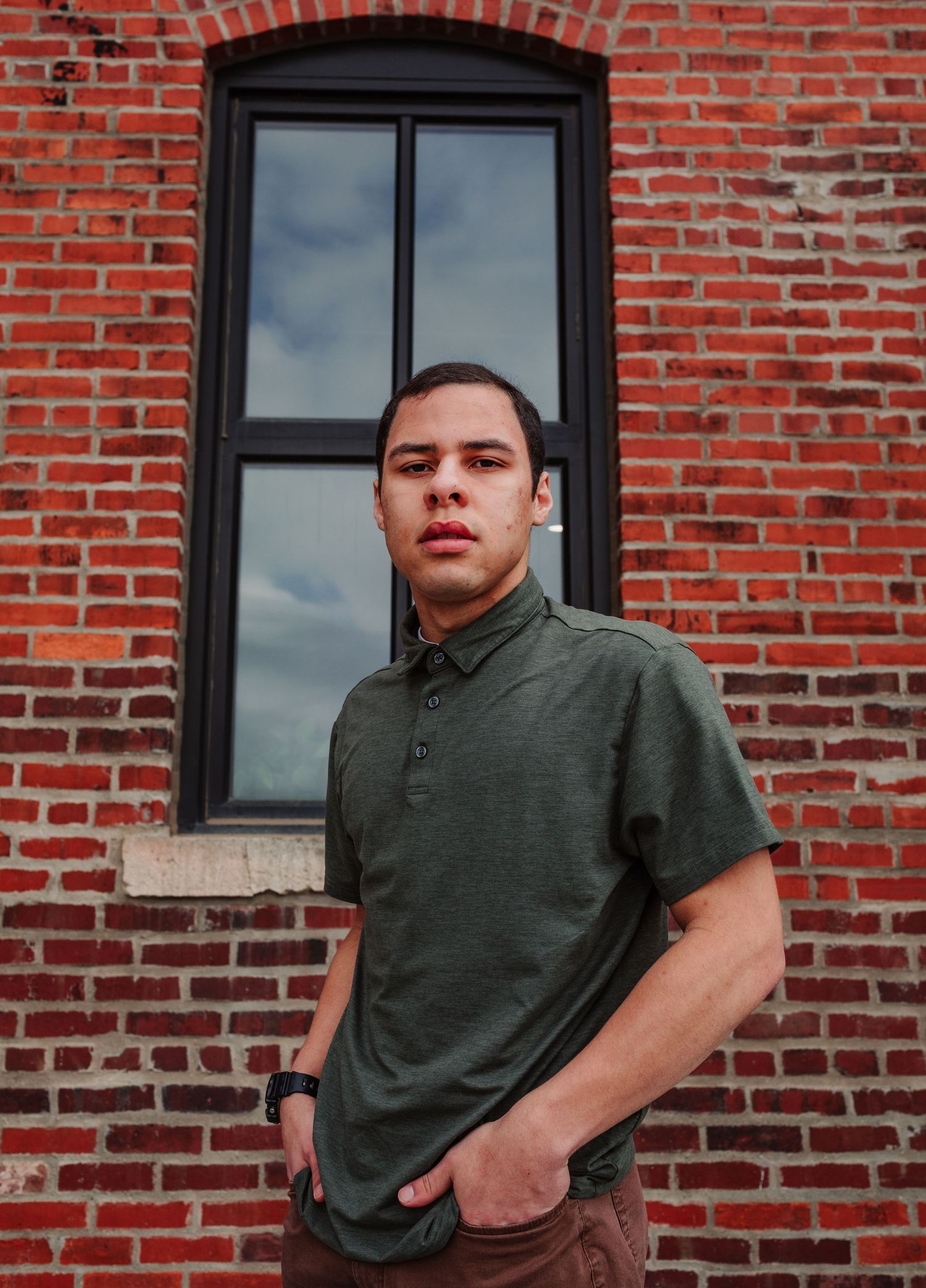 Young man in green polo shirt standing against red brick wall outdoors.