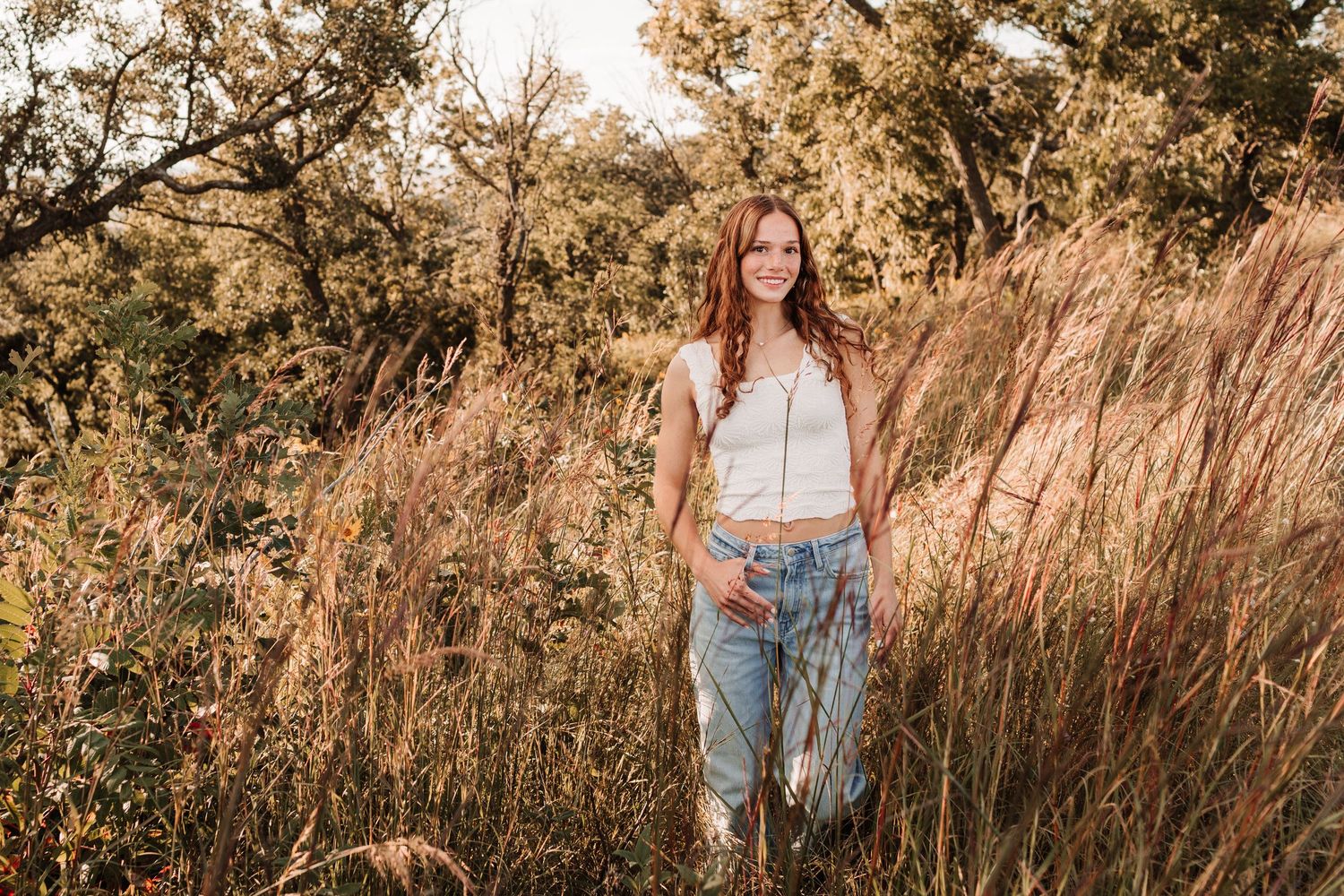 Smiling young redhead woman in white top and jeans standing among tall golden grasses in autumn field.