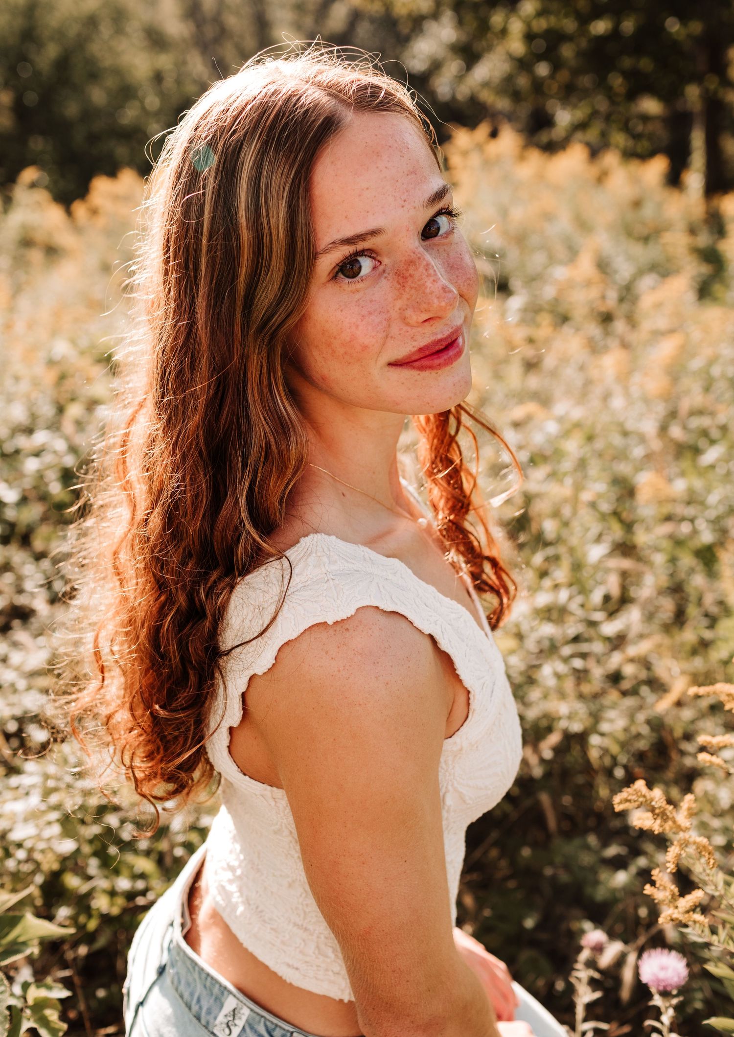 Portrait of a young freckled woman with auburn hair in white top smiling in a golden wildflower field.