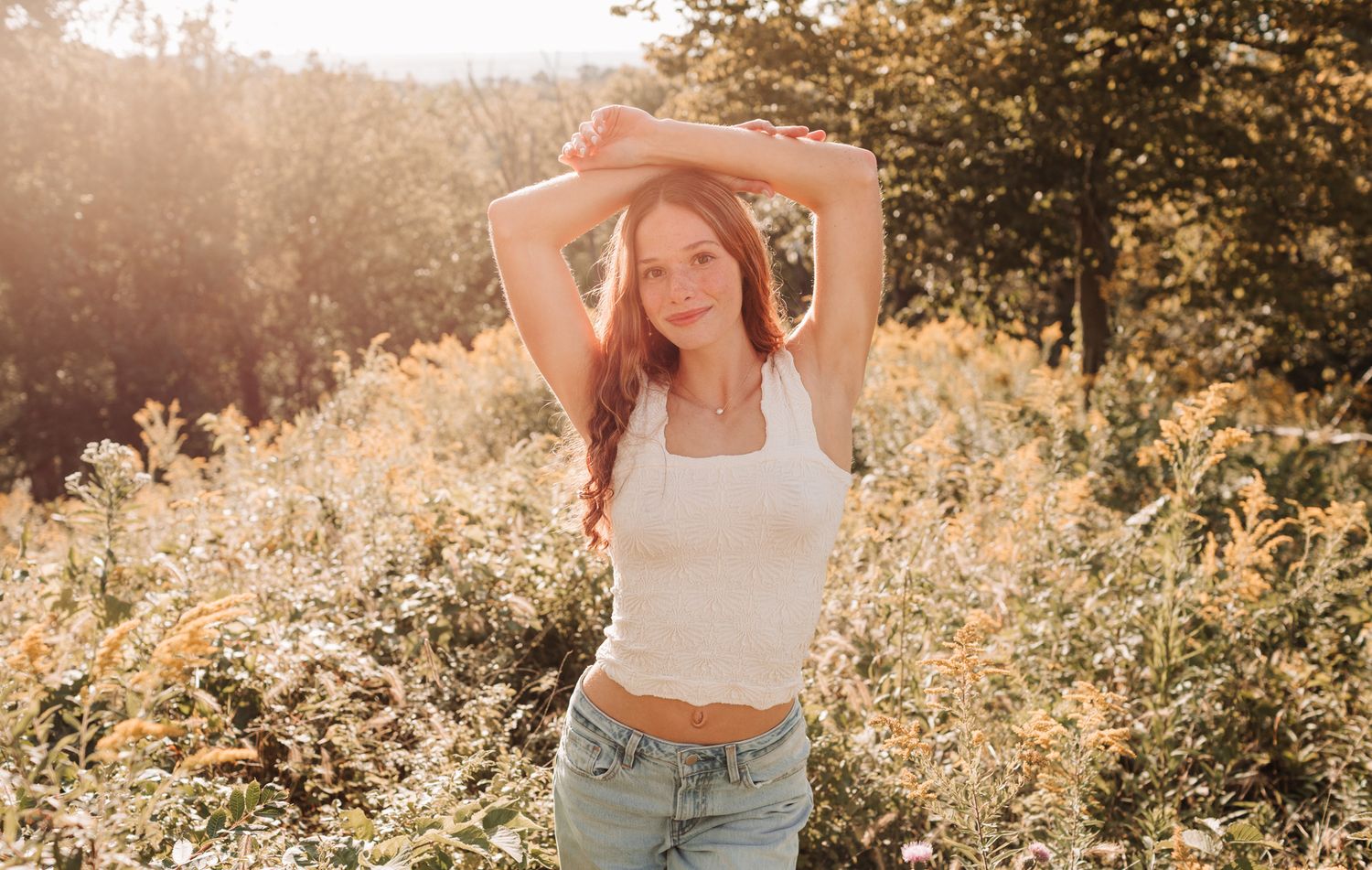 Young woman with long red hair in white crop top poses with arms raised in a bright sunny meadow.