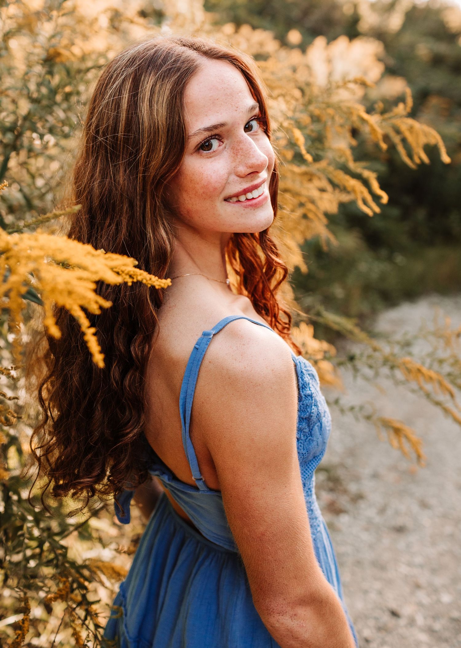 Young woman with long auburn hair in blue dress smiling over shoulder beside golden wildflowers outdoors.