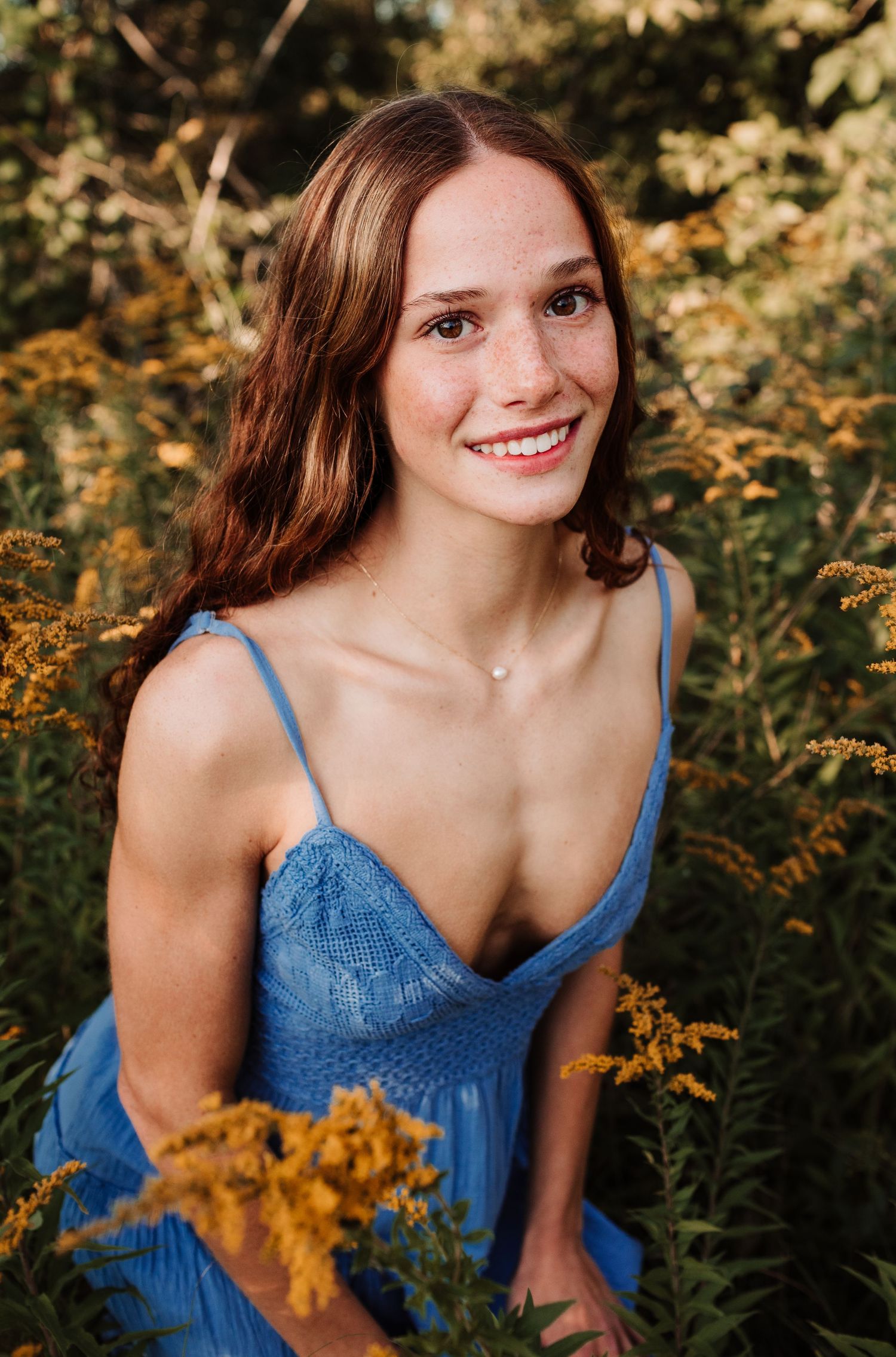 Young freckled woman with auburn hair in blue dress smiling while seated among golden wildflowers outdoors.