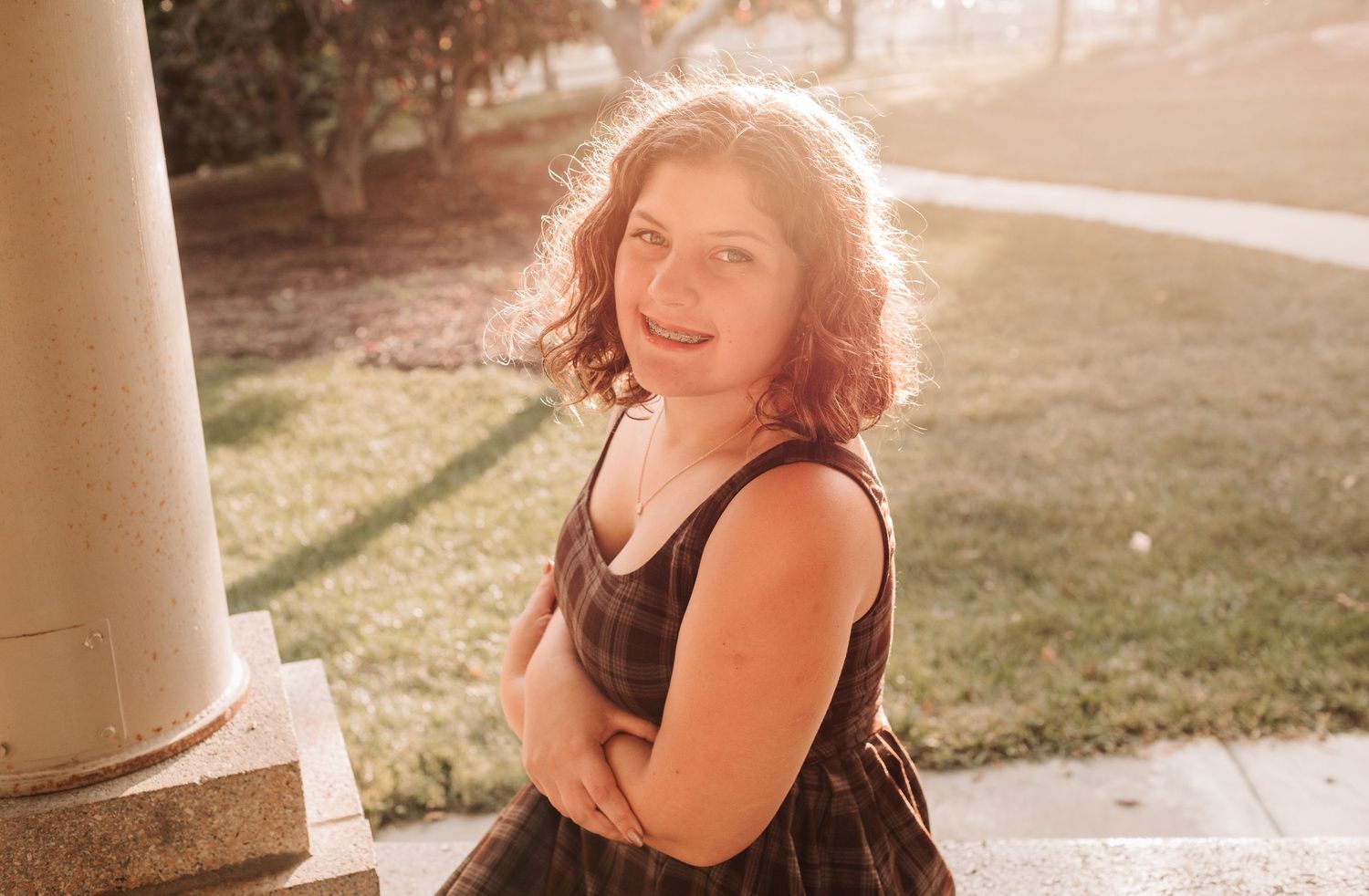 Young woman with curly hair smiling outdoors near column in golden sunlight.