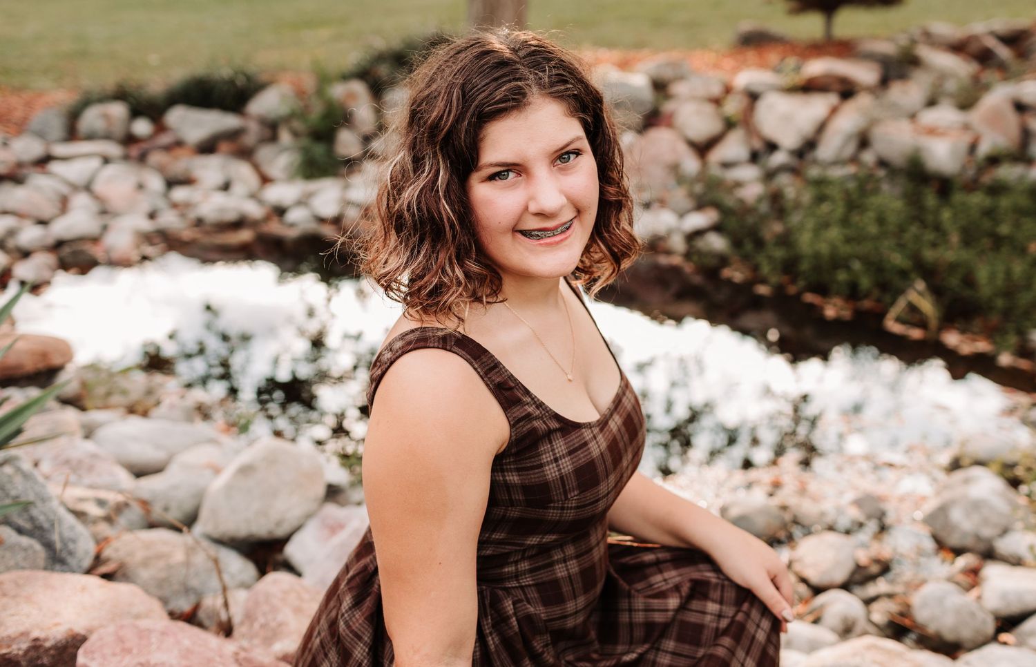 Teenage girl in plaid dress smiling near rocky pond outdoors.