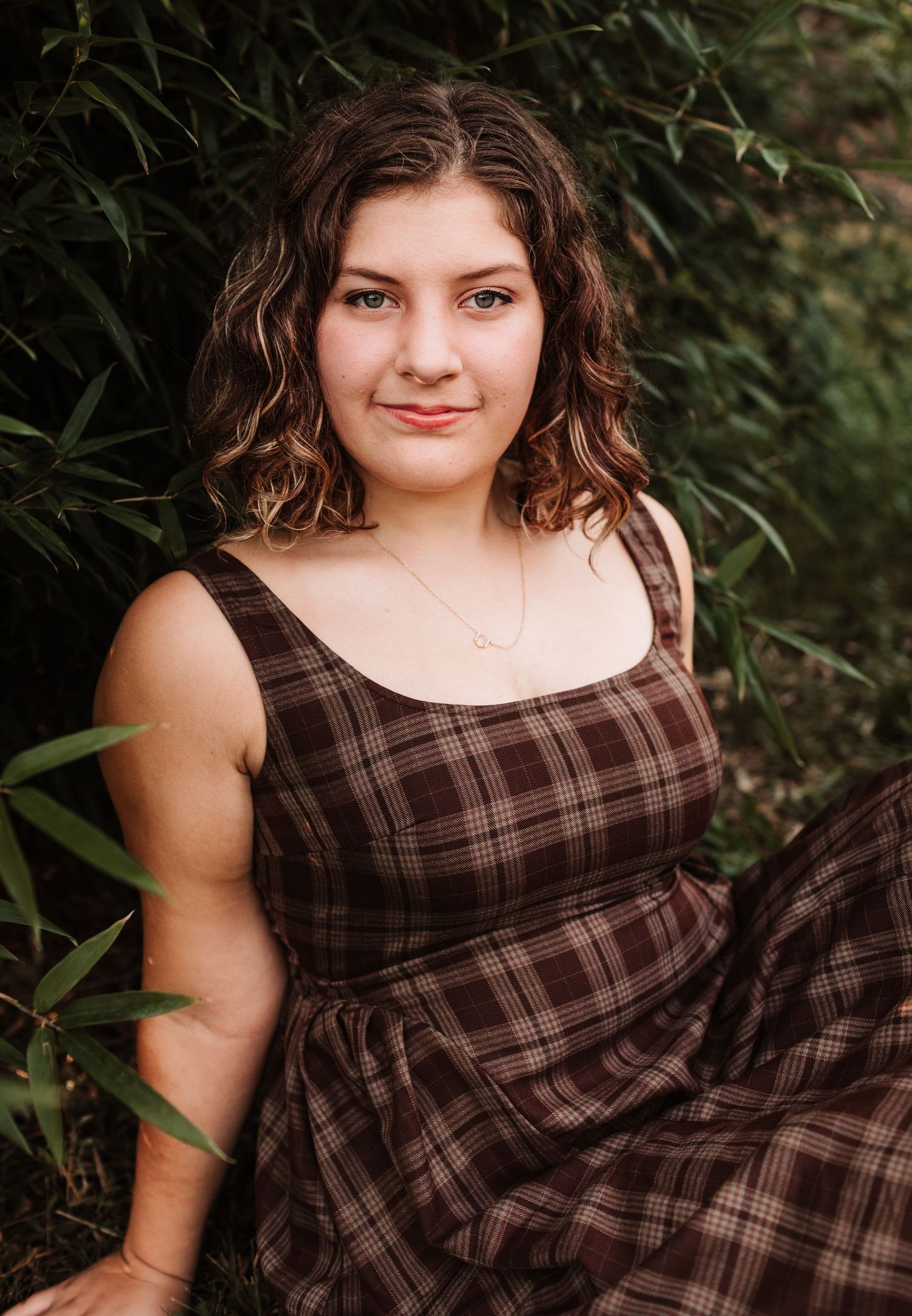 Young woman in brown plaid dress sitting among green bamboo leaves outdoors.