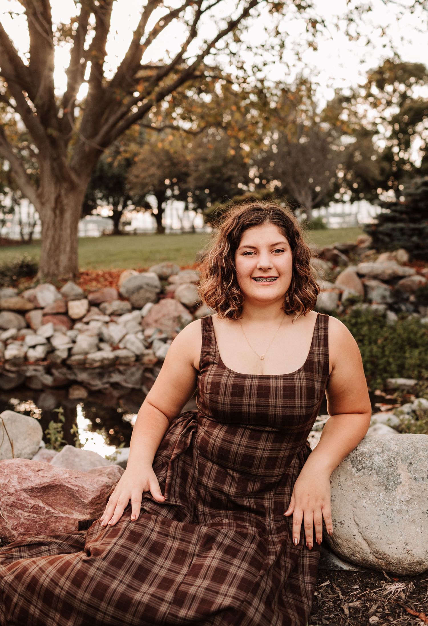 Young woman in brown plaid dress sitting on rocks near garden pond.