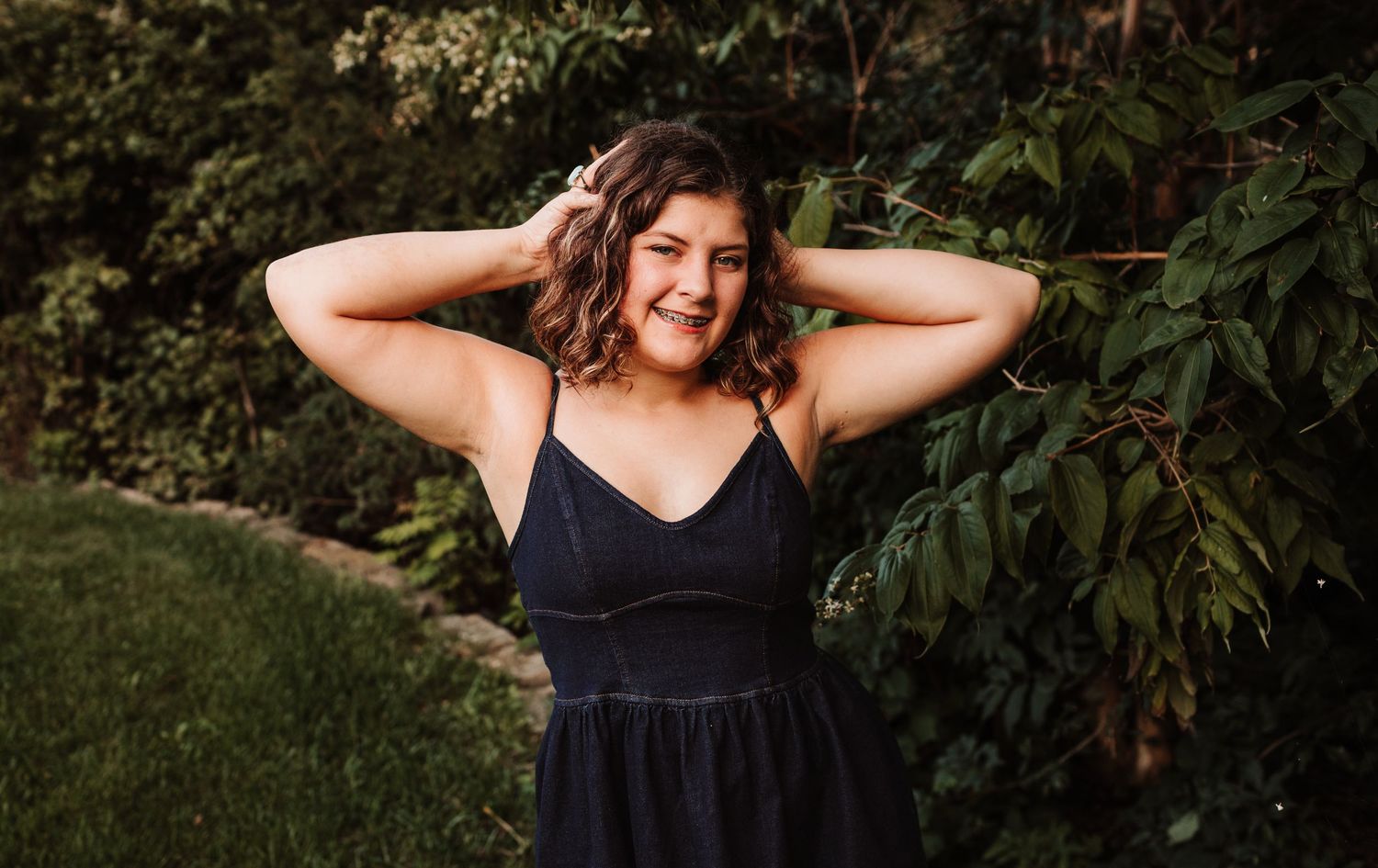 Smiling young woman in black dress posing outdoors among green foliage.