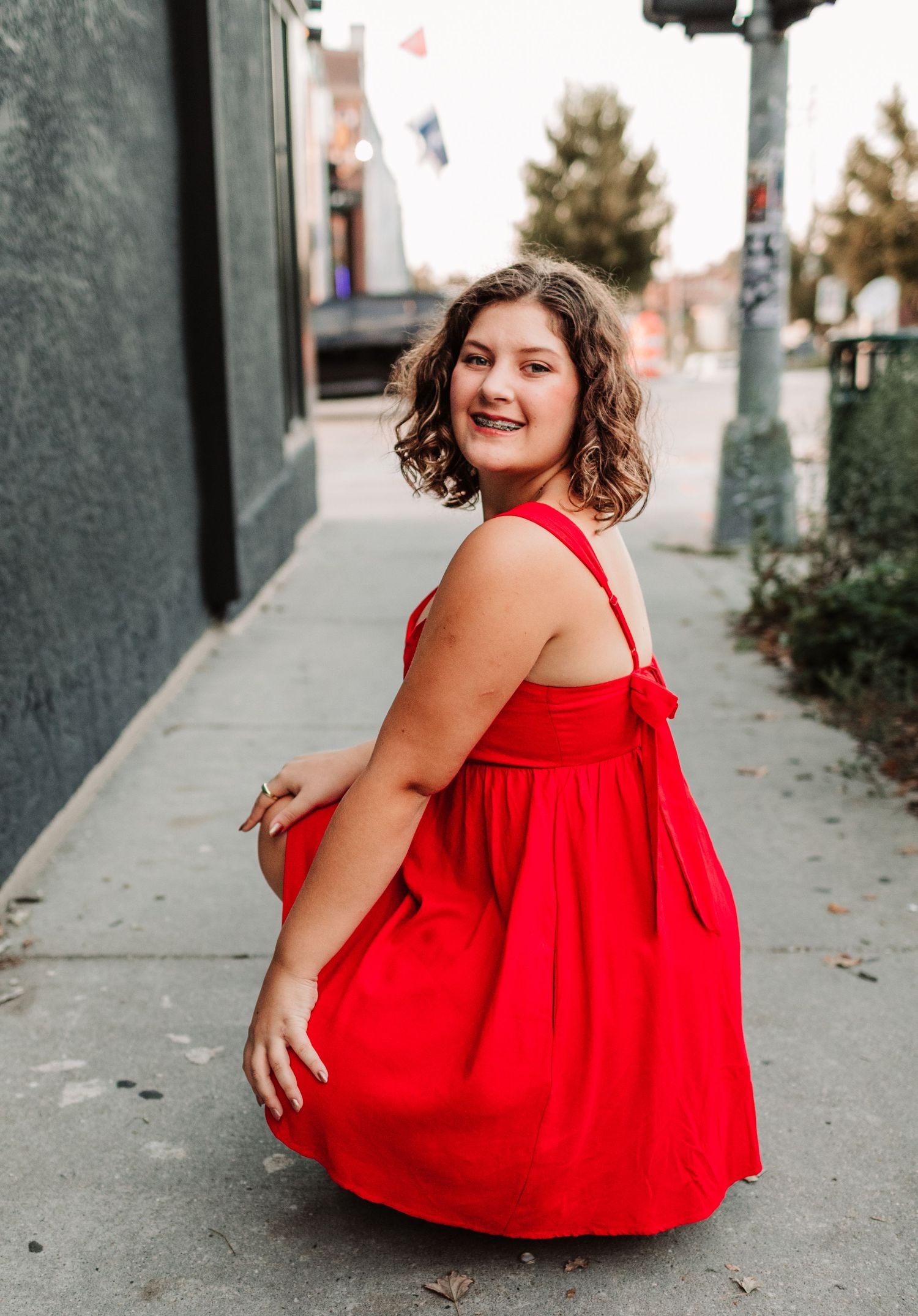 Young woman in red dress crouching on urban sidewalk smiling over shoulder.
