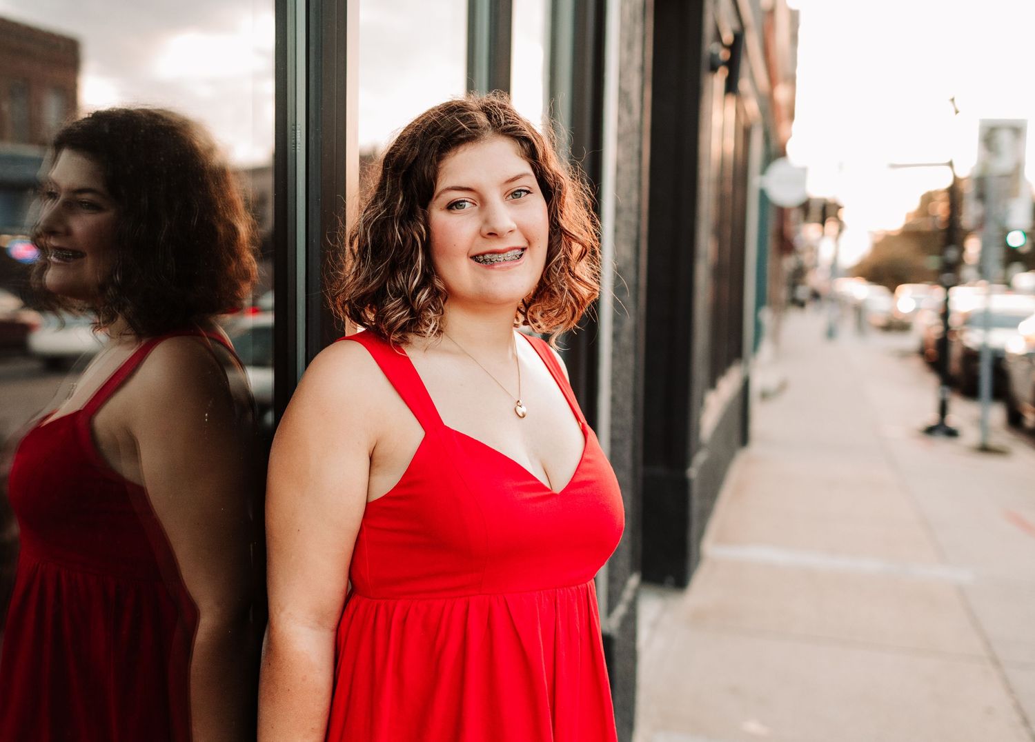 Smiling young woman in red dress leaning against glass storefront on city street.