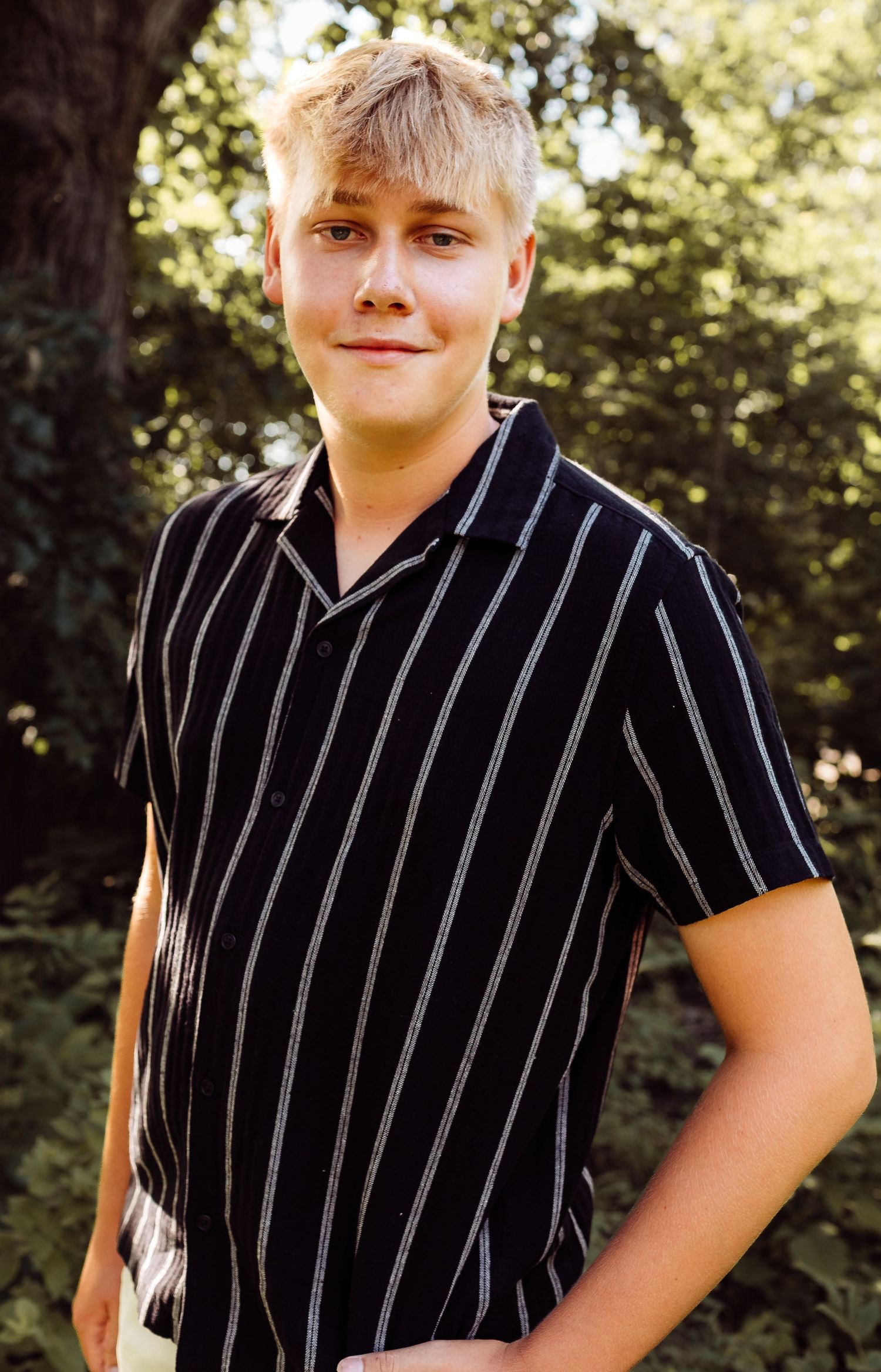 Blonde teenage boy in black striped short-sleeve shirt posing outdoors among green trees.
