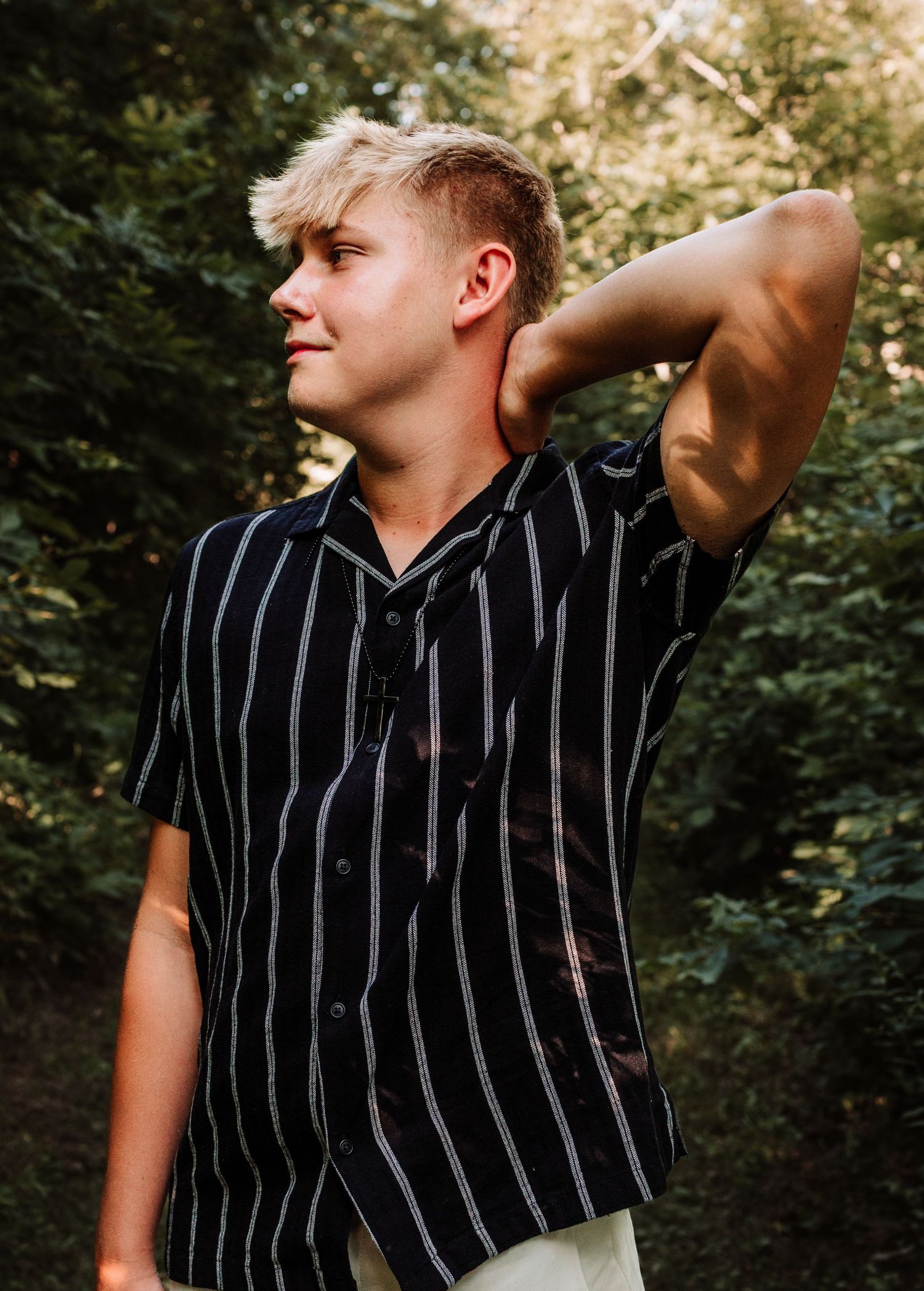 Young blonde man in striped black shirt posing with hand behind neck in sunlit forest.