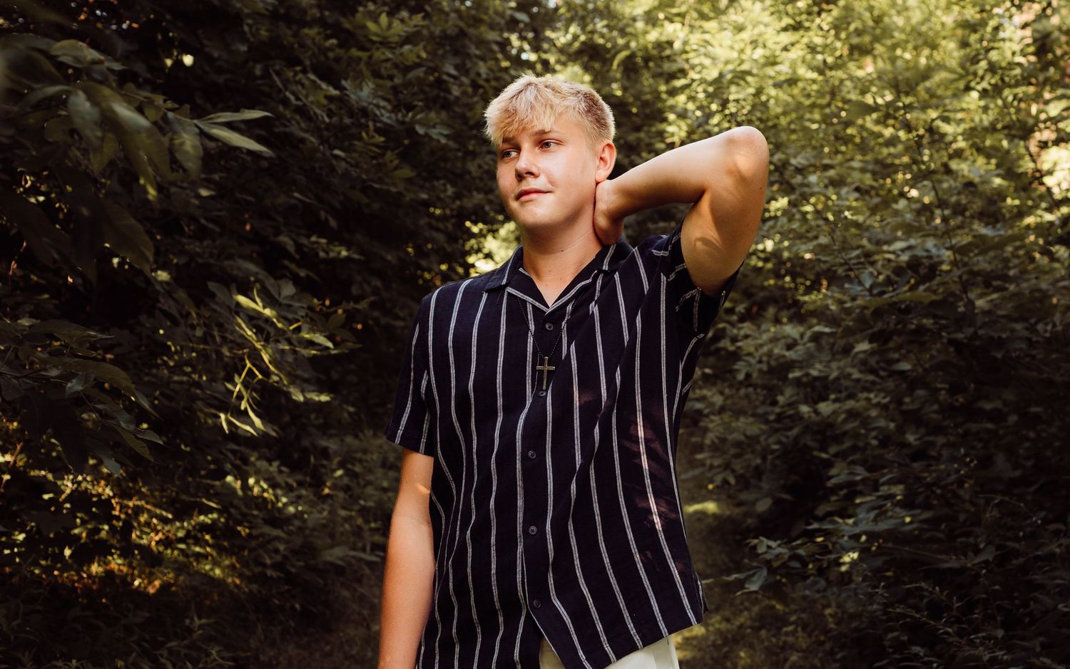 Teenage boy in black striped shirt standing confidently in a sunny green wooded area.
