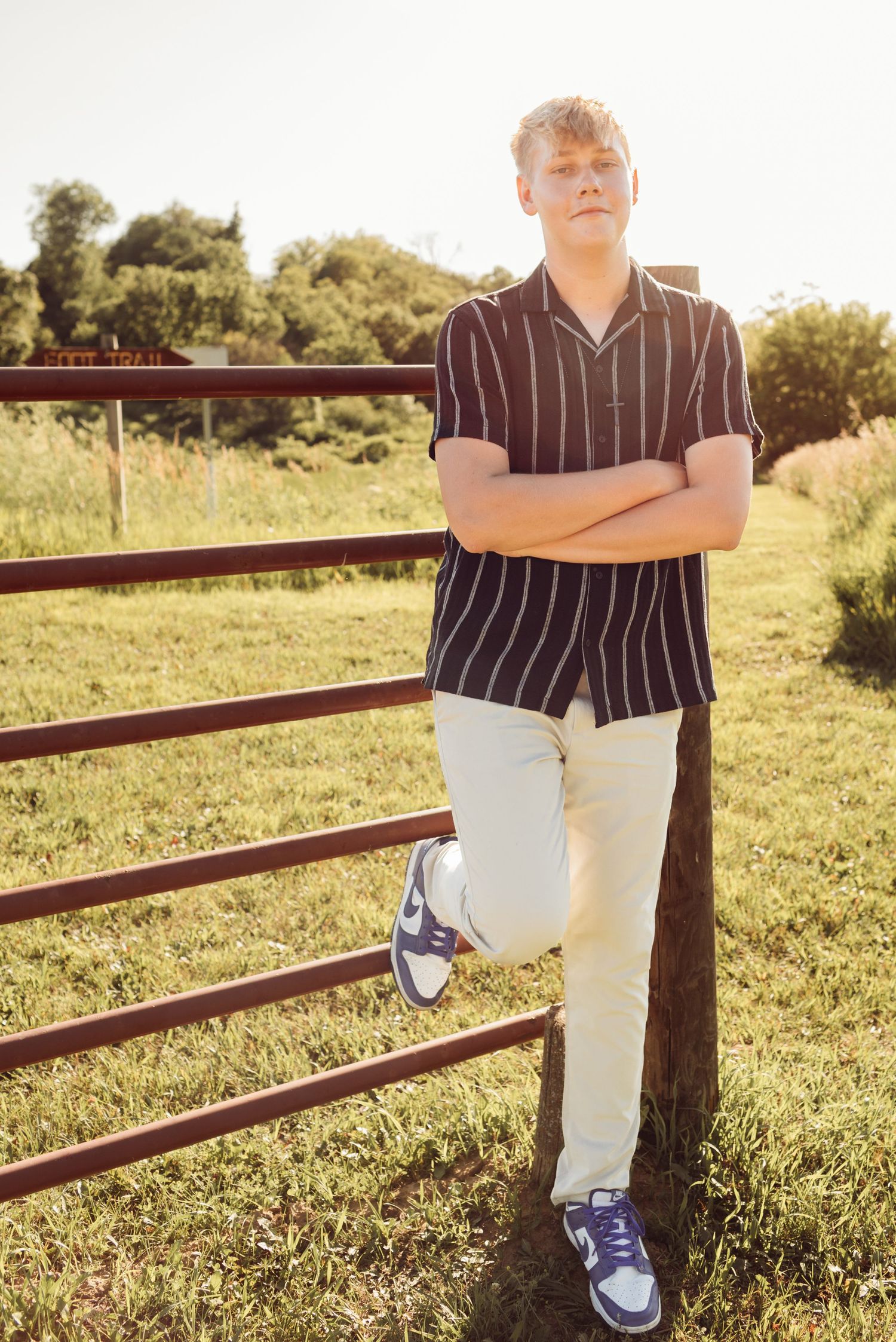 Teen boy in striped shirt and khaki pants leaning on red fence in sunny field.