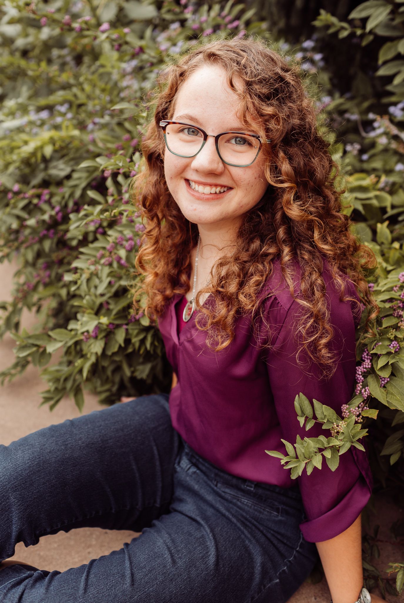 Smiling young woman with curly red hair and glasses sitting outdoors among purple flowering bushes.