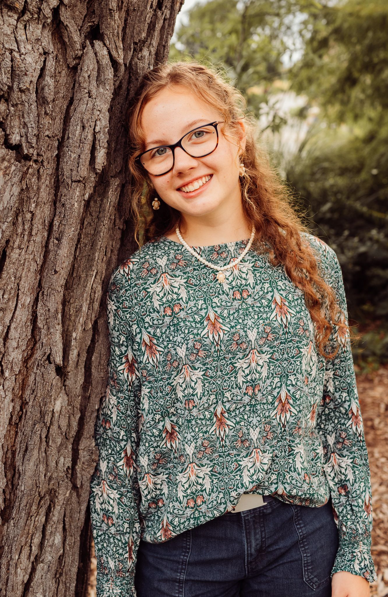 Smiling teenage girl with curly red hair and glasses leaning against a tree outdoors.