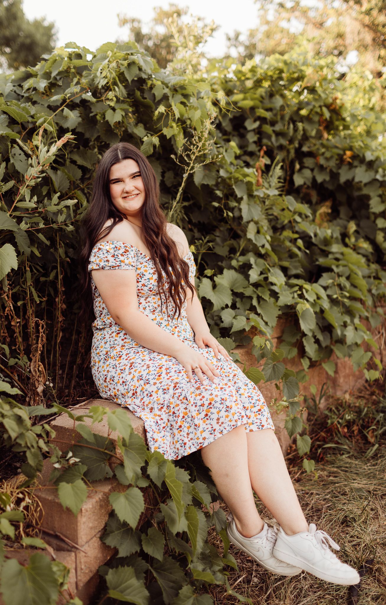 Smiling young woman in floral dress sitting outdoors surrounded by lush green foliage.