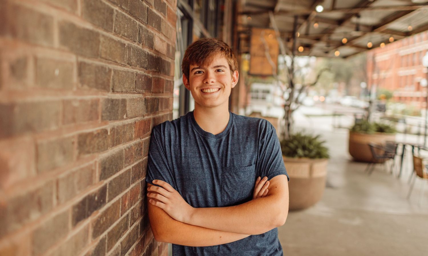 Smiling young man with arms crossed leaning against a brick wall in an outdoor setting.
