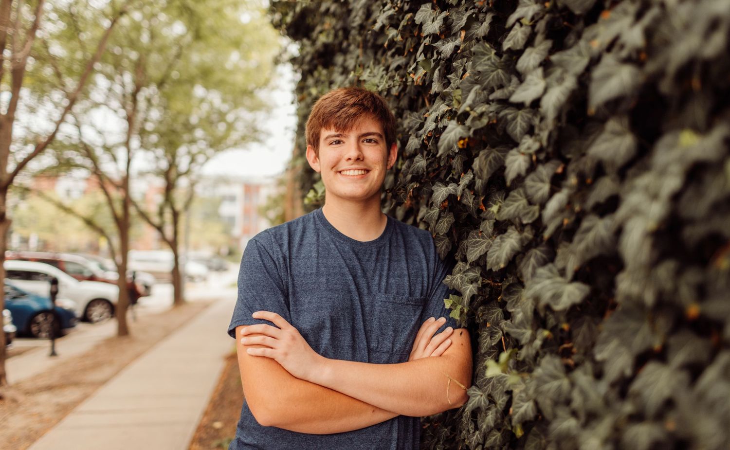 Smiling young man with arms crossed leaning against an ivy-covered wall on a city sidewalk.
