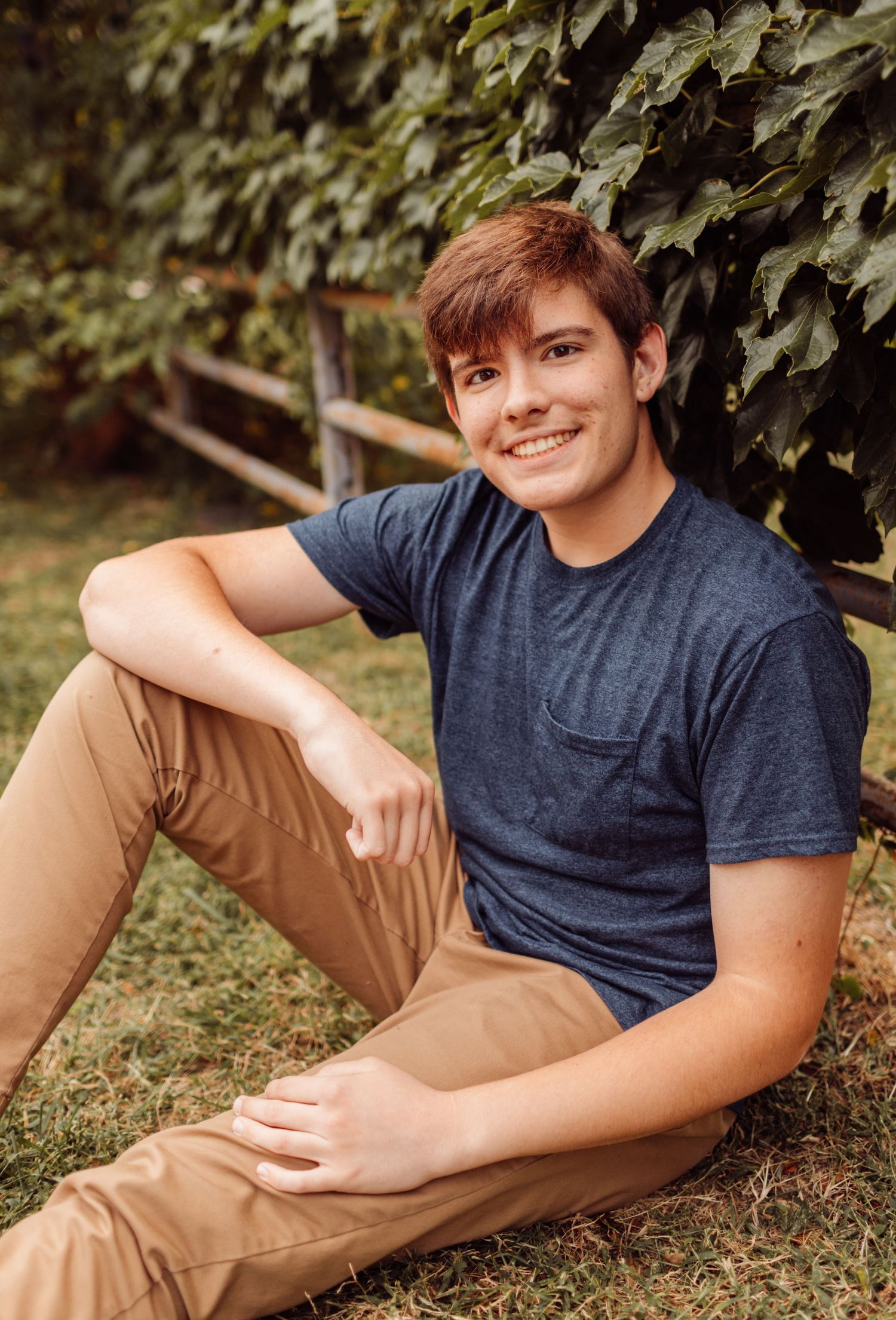 Smiling teenage boy in blue t-shirt sitting on grass in front of green ivy fence.