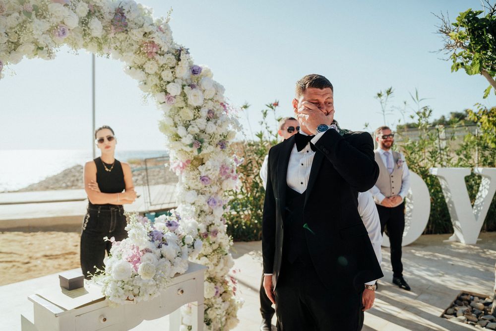 A wedding ceremony takes place under a floral arch with white flowers and greenery against a scenic outdoor backdrop.