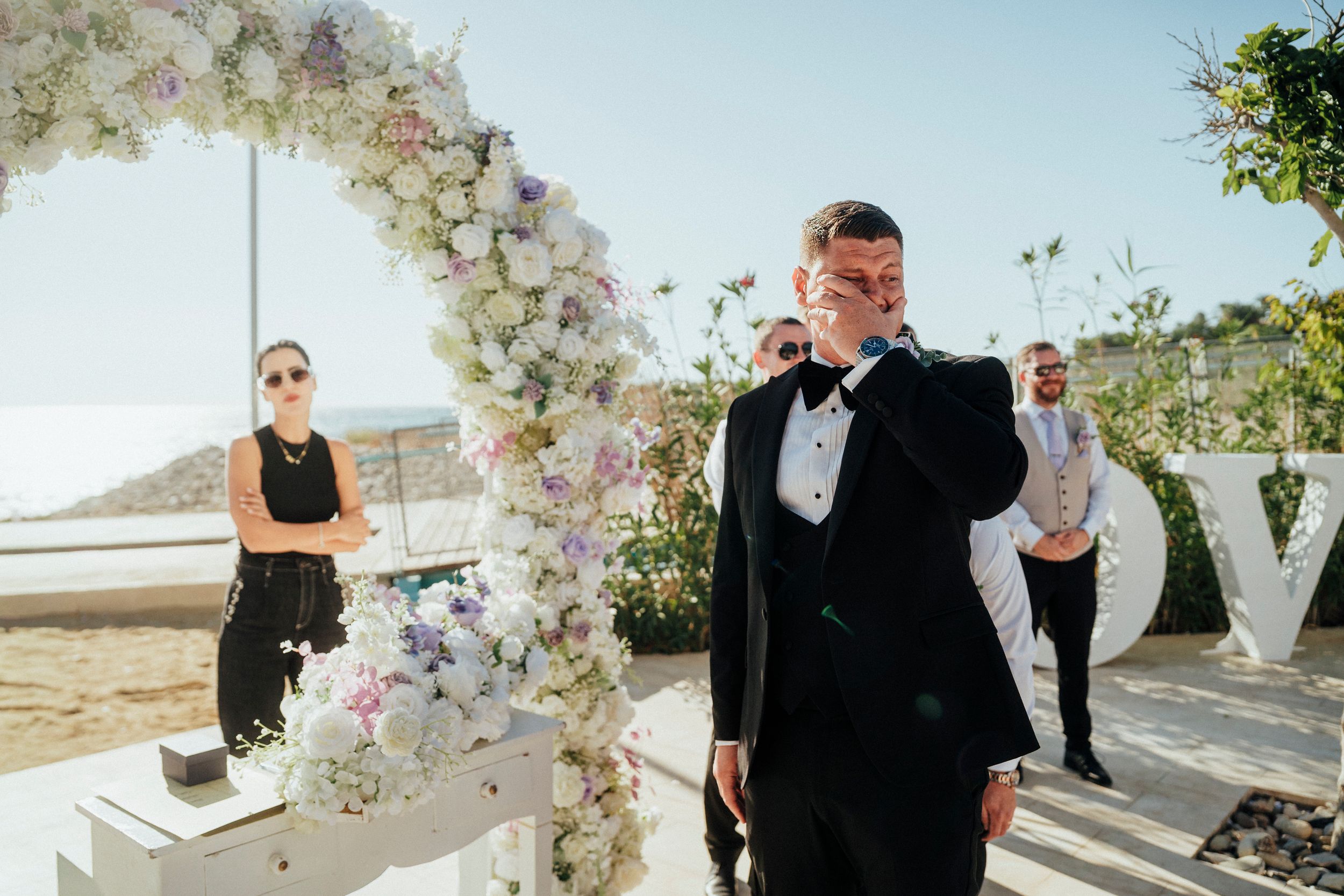 A wedding ceremony takes place under a floral arch with white flowers and greenery against a scenic outdoor backdrop.