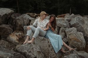 Two women sitting barefoot on large rocks outdoors, one in a hat and white shirt, the other in a blue skirt, smiling together.