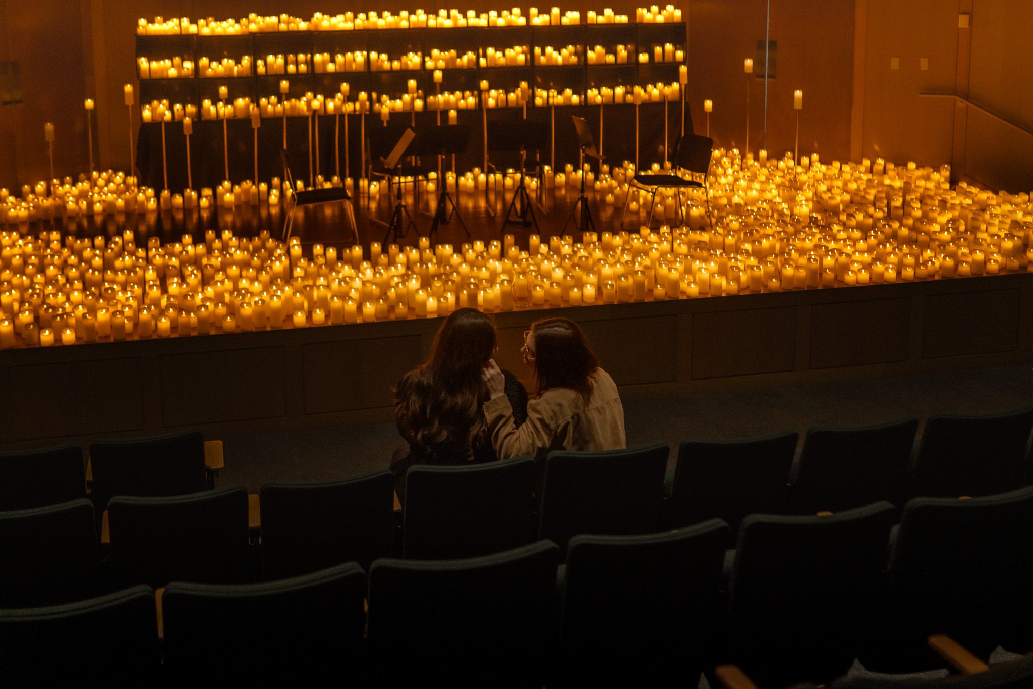 Rows of glowing candles illuminate a dark theater stage with empty seats in the foreground.