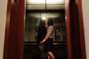 Two people standing in a dimly lit elevator with reflective metal walls and a glowing ceiling panel.