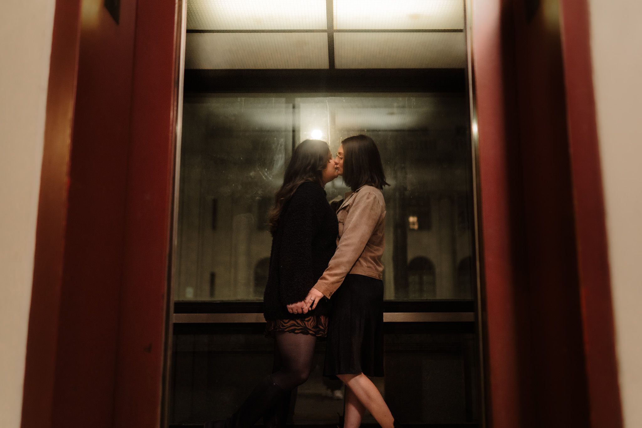 Two people standing in a dimly lit elevator with reflective metal walls and a glowing ceiling panel.