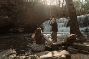 Couple near a sunlit waterfall on rocky riverbank, man photographing woman with long hair in gray dress outdoors.