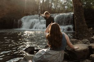 Couple sitting by a sunlit waterfall on rocky riverbank surrounded by lush trees in a romantic outdoor setting.