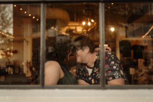 Couple sharing a romantic kiss through a window with warm bokeh lights in the background.