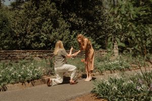 Person proposing on one knee to partner in a lush garden setting with stone wall and blooming flowers.