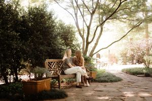 Two women sitting together on a wooden bench in a lush garden surrounded by green trees and plants.